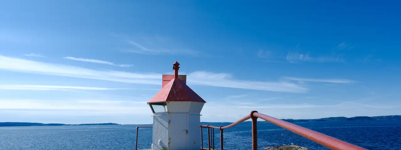 Lite hvitt fyr med rødt tak på et svaberg ved Tofte, med rekkverk langs berget og Oslofjorden bak under klar himmel.