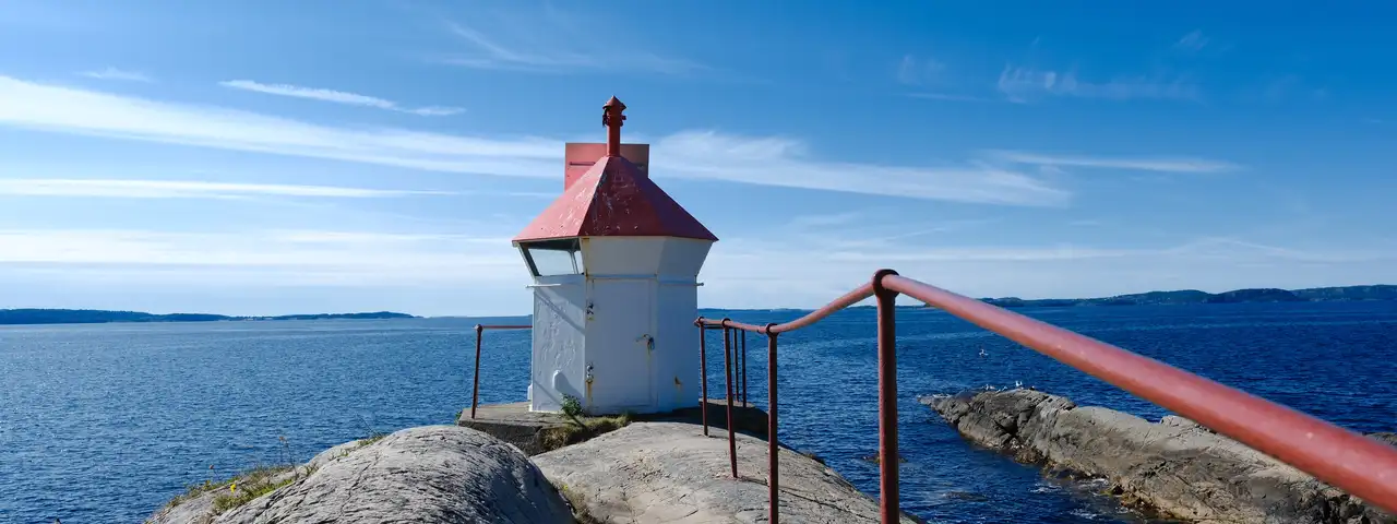 Liten hvit fyrlykt med rødt tak på et svaberg ved Tofte, med rødt rekkverk i forgrunnen og fjorden bak.