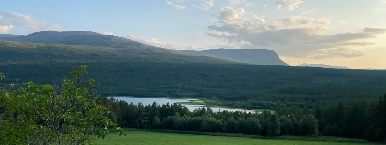 Grønt jorde med gjerdestolpe i forgrunnen, skog og et vann under, og et bredt fjell i kveldslys bak.