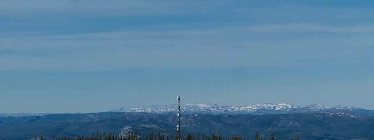 Mobilmast som stikker opp over granskogen på en høyde, med blåner og snødekte fjell i bakgrunnen.