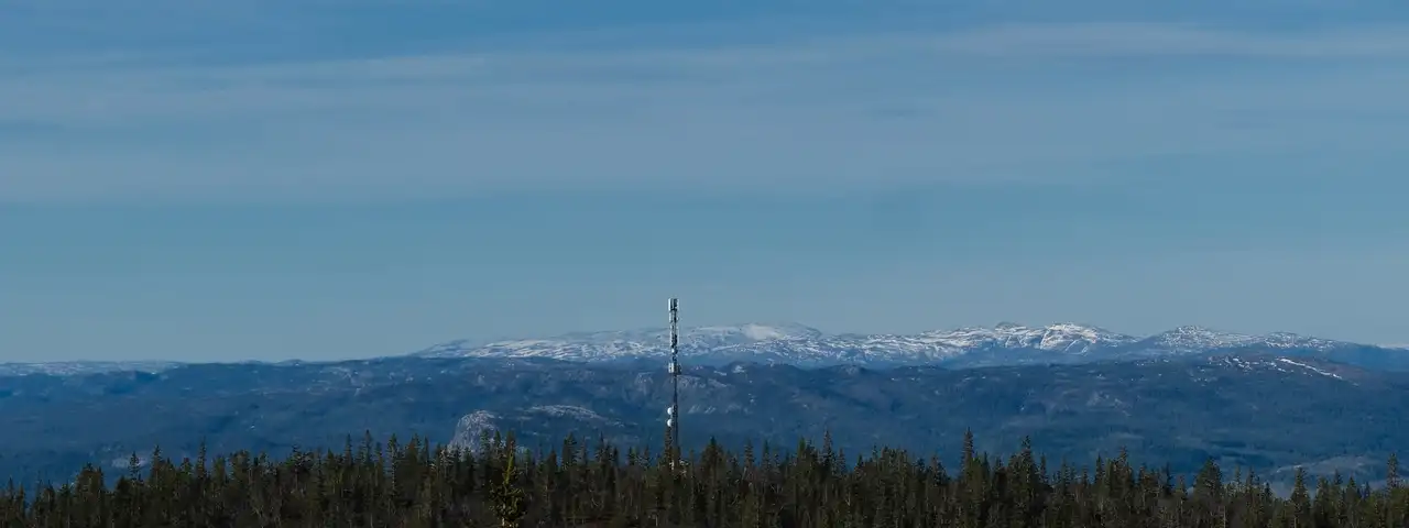 Mobilmast som stikker opp av skogen på en åskam, med snødekte fjell i horisonten.