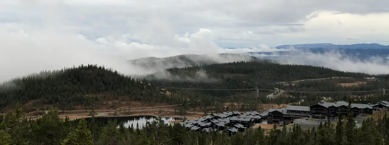 Lavt skydekke som driver inn over skogkledde åser, med en klynge mørke hytter og et større byggverk ved et lite vann.
