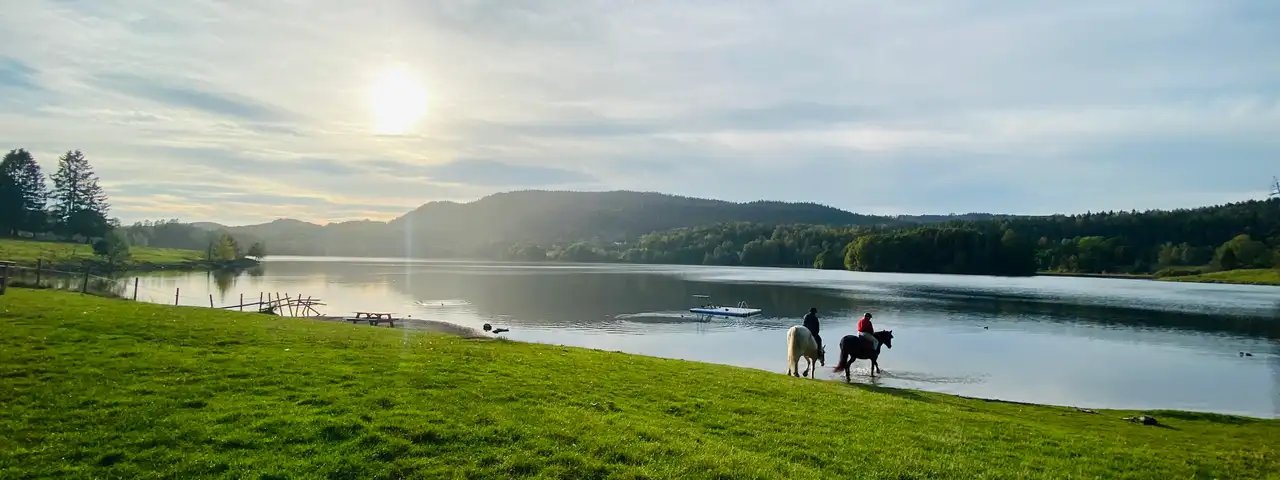 To ryttere til hest i vannkanten av Semsvannet, med lav sol over åsene bak.