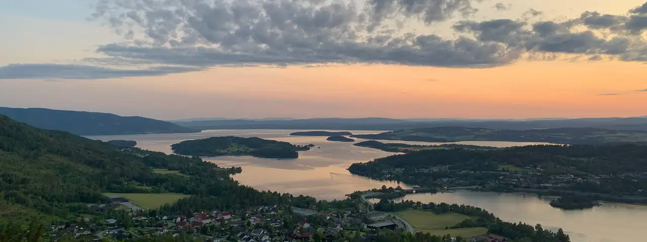 Panorama over Sundvollen i solnedgang, med boligfelt og skog i forgrunnen og Tyrifjordens øyer og åser bak under oransje kveldshimmel.
